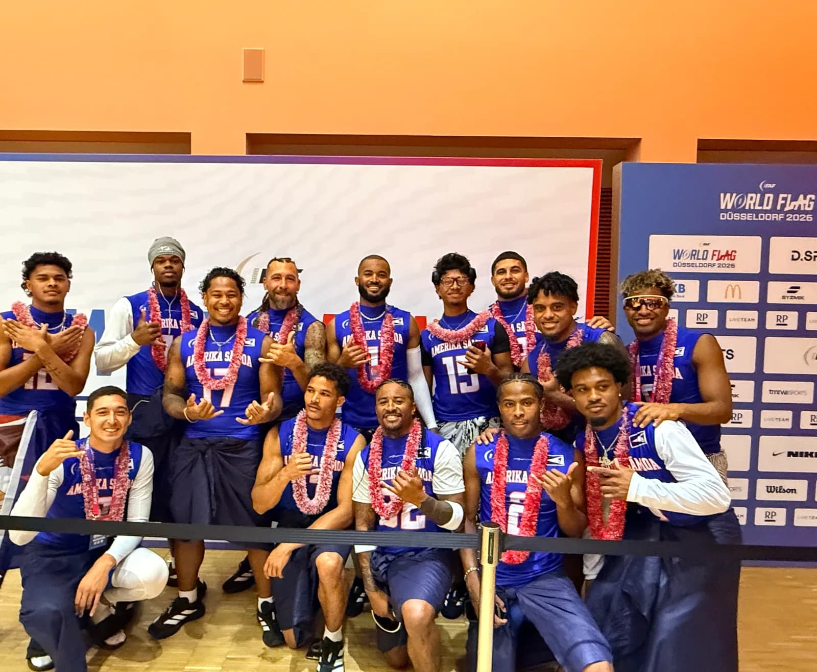 The squad in blue Amerika Sāmoa jerseys wearing ʻula, throwing shakas in front of the IFAF World Flag Düsseldorf 2026 backdrop.