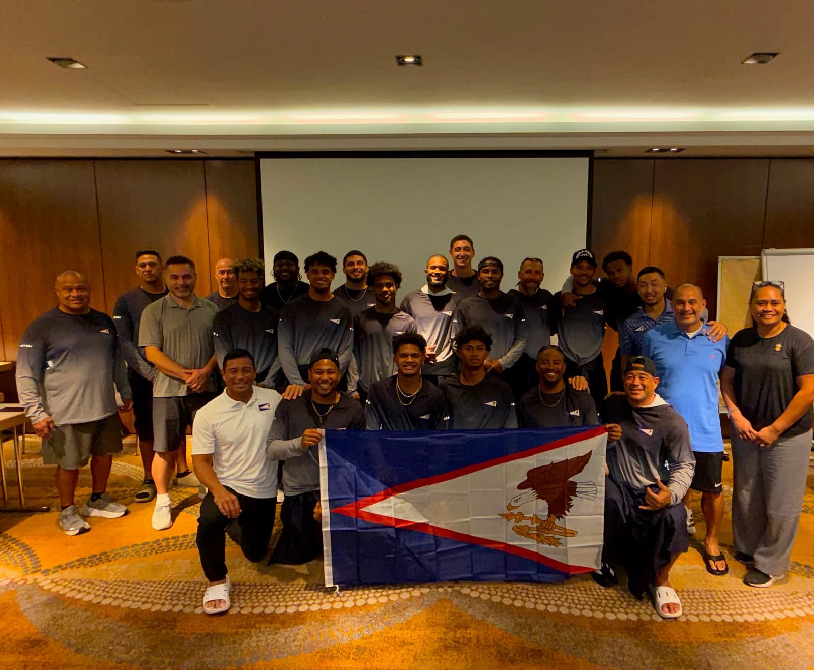 The full travelling party, players, coaches and staff, standing together indoors behind the flag of American Samoa.