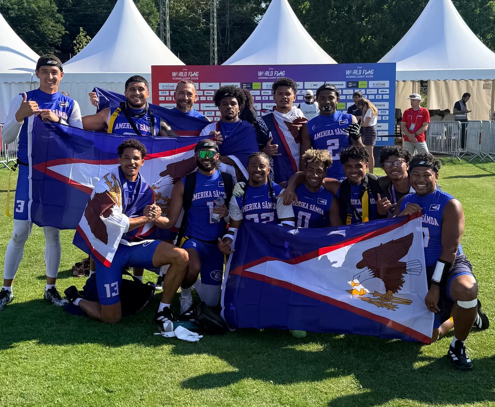 The American Samoa squad on the field in Düsseldorf, holding two American Samoa flags in front of the IFAF World Flag Düsseldorf 2026 backdrop.